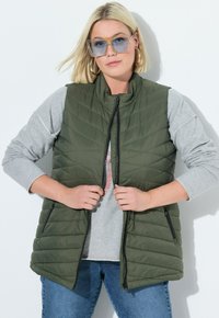 Olivenfarbene gesteppte Weste mit Reißverschluss vorne, seitlichen Taschen und hohem Kragen, kombiniert mit einem grauen Langarmshirt und Jeans.
