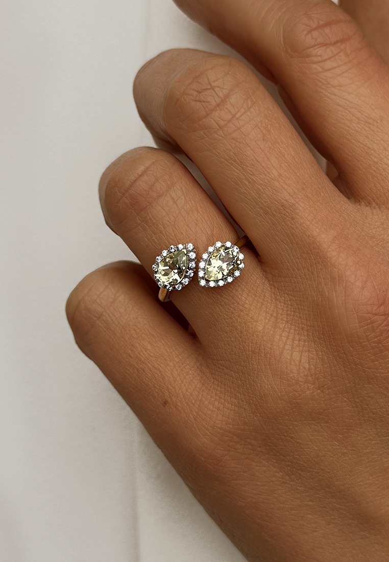 Bague en or ornée de deux cristaux en forme de poire entourés de petites pierres transparentes. Présentée à la main sur un fond clair.