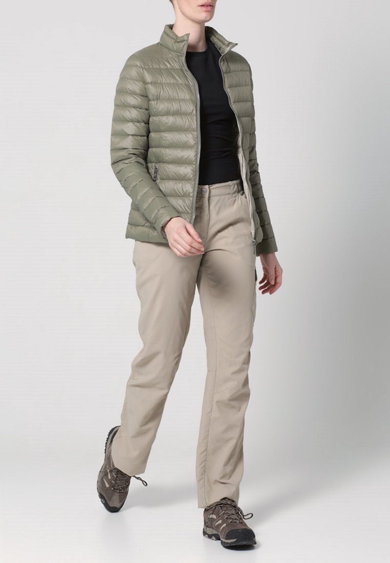 Femme marchant portant une doudoune vert olive clair, un haut noir, un pantalon beige et des chaussures de randonnée marron sur un fond gris uni.