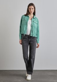 Veste courte teal en tissu doux avec deux poches avant et un ourlet à cordon, associée à un jean noir et des baskets blanches.