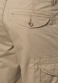 Shorts cargo beige avec poches à rabat boutonnées à l'arrière et sur les côtés, montrant des coutures détaillées et la texture du tissu.