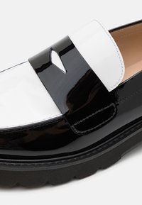 Mocassin en cuir verni noir et blanc avec une semelle noire épaisse et une bride noire distinctive comportant un petit détail découpé sur le dessus.