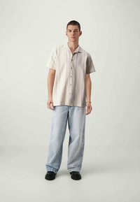 Fiatalember áll, bézs színű, rövid ujjú, gombos inget visel, világoskék, bő nadrágot, fekete cipőt és egy karkötőt a jobb csuklóján.