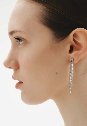 Perfil de una mujer de piel clara y cabello oscuro que lleva pendientes largos, plateados y rectangulares, sobre un fondo blanco liso.