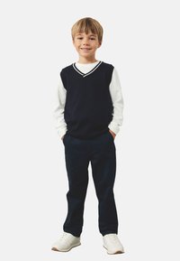 Marineblauwe gebreide vest met witte afwerking over een wit shirt met lange mouwen, gecombineerd met donkere broek en witte sneakers. Lachend kind poseert.