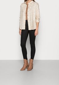 Kvinna som bär en beige skjorta med djurtryck, brun topp, svarta tajta byxor och tanfärgade spetsiga ankelstövlar med klack.