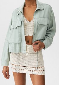 Veste courte vert clair avec deux poches avant, portée sur un haut blanc en crochet et une jupe assortie présentant des motifs complexes.