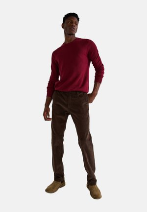 Pull à col rond rouge, pantalon en velours côtelé marron et bottines chevilles beige clair. Le modèle pose avec les bras détendus, mettant en valeur une coupe ajustée et une texture.