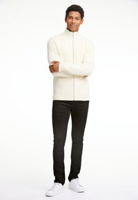 Suéter de cremallera color crema con textura acanalada, combinado con jeans ajustados negros y zapatillas blancas. El modelo está de pie con los brazos cruzados frente a un fondo blanco.