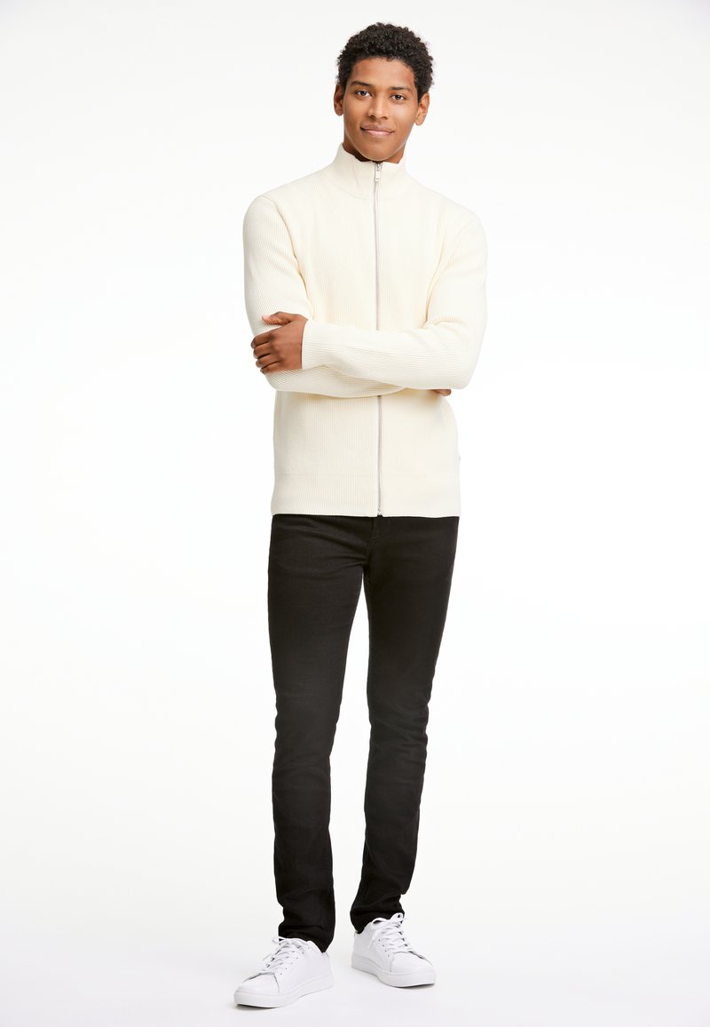 Suéter de cremallera color crema con textura acanalada, combinado con jeans ajustados negros y zapatillas blancas. El modelo está de pie con los brazos cruzados frente a un fondo blanco.