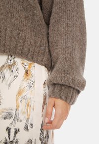 Brauner, gestrickter Pullover mit gerippten Bündchen, kombiniert mit einem cremefarbenen Rock, der detaillierte Illustrationen verschiedener Tiere zeigt.