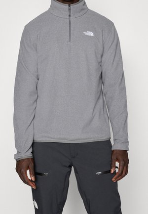 Mężczyzna w szarym polarze z krótkim suwakiem i logo The North Face oraz czarnych spodniach z kieszeniami zapinanymi na zamek, stojący na jednolitym tle.