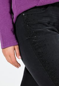 Jeans negros con un lado texturizado que presenta un patrón de pequeños goterones en relieve. La tela principal es suave y tiene un ligero brillo.