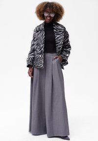 Zwart-witte jas met zebraprint en grote knopen, gedragen over een zwarte top en hoge grijze plooibroek, gecombineerd met oversized zonnebril.