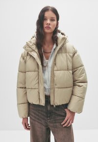 Giacca puffa beige con design corto, grande cappuccio e texture trapuntata. Abbinata a un felpa grigia e jeans scuri a vita alta.