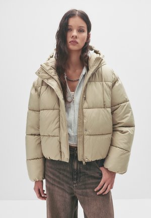 Jachetă puffer bej cu un design scurt, glugă mare și textură matlasată. Asortată cu un hanorac gri și blugi închiși la culoare, cu talie înaltă.