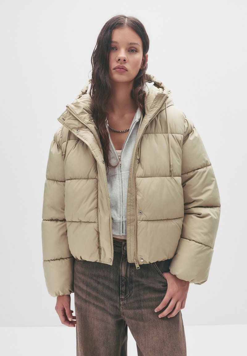 Giacca puffa beige con design corto, grande cappuccio e texture trapuntata. Abbinata a un felpa grigia e jeans scuri a vita alta.