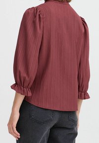 Bordeauxbluse med tekstureret stof, samlet pufærmer og flæsekant. Har en simpel krave og lige kant. Båret med mørke jeans.