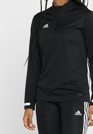 Frau trägt ein schwarzes Adidas Langarmshirt und dazu passende schwarze Adidas Trainingshose mit weißen Streifen und Logos.