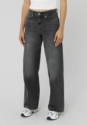 Femme portant un jean noir délavé taille haute à jambes larges, des baskets blanches et un haut blanc sans manches, debout devant un fond gris.