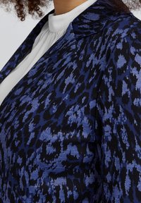 Blazer con stampa leopardata blu e nera realizzato in tessuto testurizzato, con colletto e camicia bianca liscia sotto.