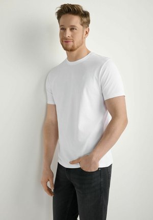 Młody mężczyzna z jasnobrązowymi włosami i brodą, ubrany w prostą białą koszulkę i czarne dżinsy, stoi oparty o białą ścianę, z ręką w kieszeni.