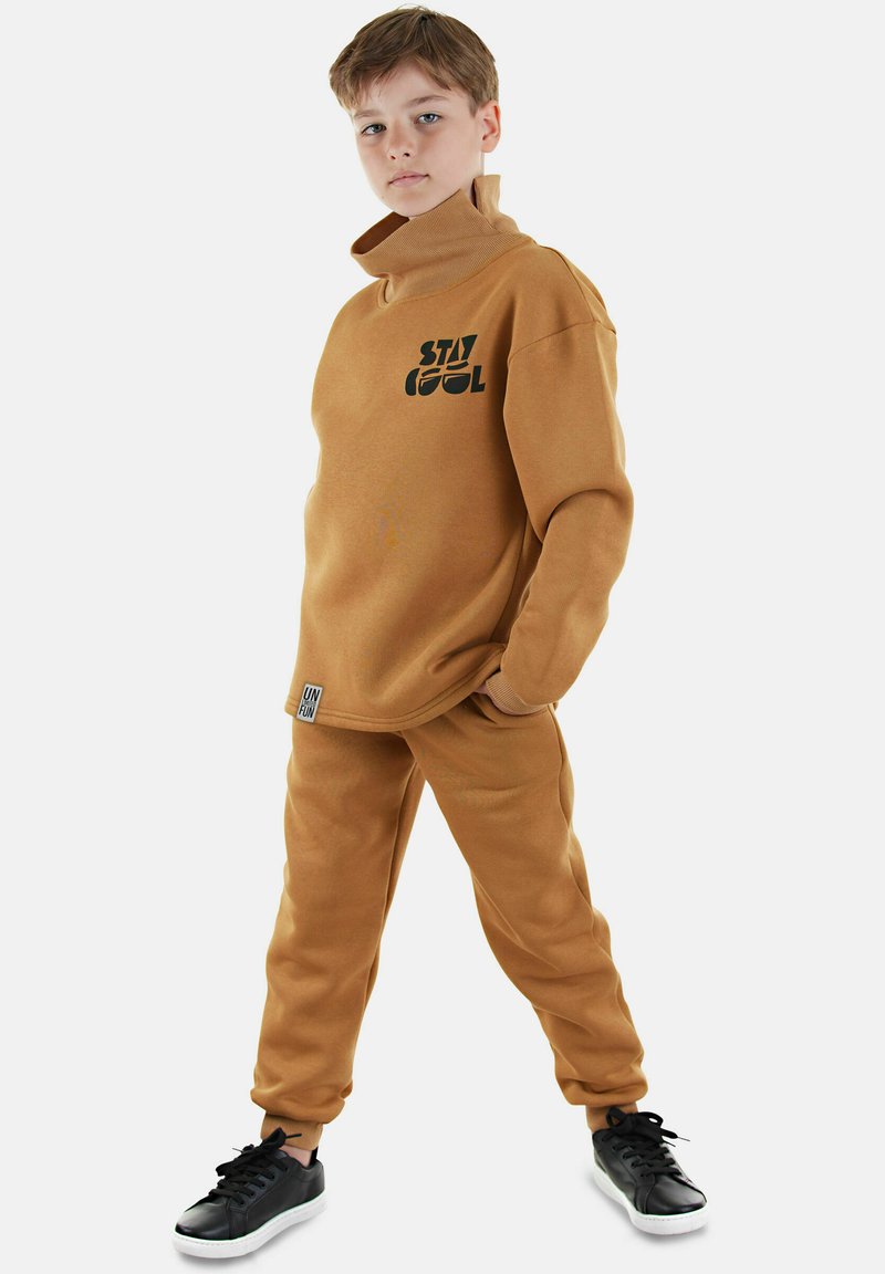 Brun sweatshirt och matchande byxor av mjukt material. Har hög krage, svart "STAY COOL" tryck och "UNFUN" etikett på byxorna. Svarta sneakers.
