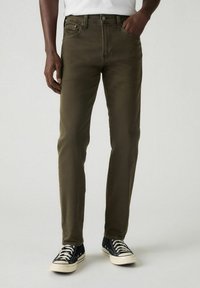 Mężczyzna w oliwkach slim-fit i czarnych niskich trampkach stoi z jedn�ą ręką w kieszeni.