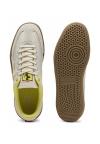 Puma-Ferrari sneakers har en overdel i beige ruskind, gule detaljer og en brun gummisål med cirkulære mønstre. Ferrari-logoet er på tungen.