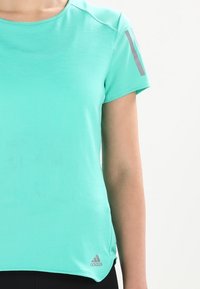 Una maglietta atletica turchese realizzata in un tessuto strutturato, con maniche corte e dettagli grigi sulle spalle. Logo Adidas sull'orlo.