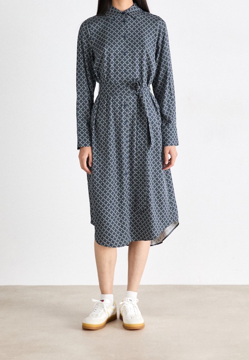 Femme portant une robe bleu marine à manches longues boutonnée, avec un motif géométrique blanc, taille nouée, chaussettes blanches et baskets blanches.