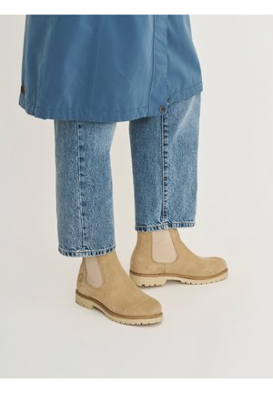 Beige suède enkellaarsjes met elastische zijpanelen, gedragen met lichtblauwe rechte jeans en een blauwe mantelzoom.