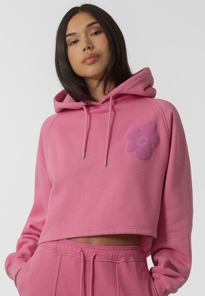 Sweat à capuche court rose avec capuche et cordons de serrage. Présente un appliqué en forme de fleur sur la poitrine. Tissu doux au toucher lisse.