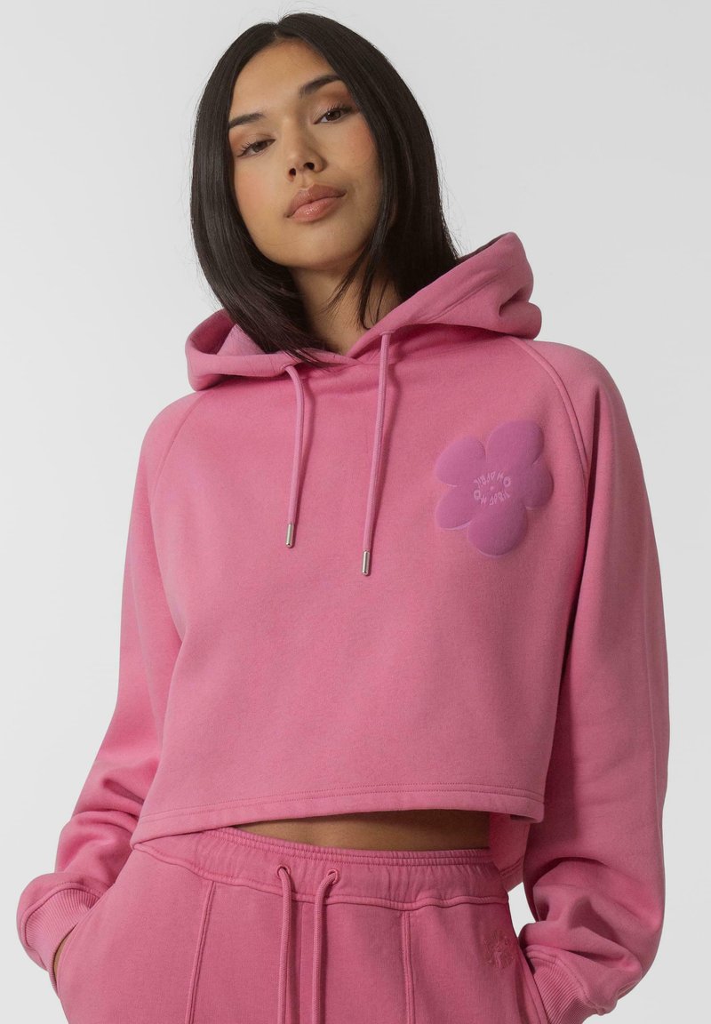 Sweat à capuche court rose avec capuche et cordons de serrage. Présente un appliqué en forme de fleur sur la poitrine. Tissu doux au toucher lisse.