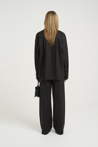 Camicia nera a maniche lunghe con polsini a bottoni, abbinata a pantaloni neri a gamba larga. La figura tiene una piccola borsa nera. Texture liscia, design minimale.