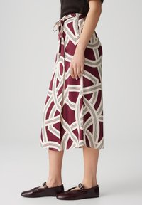Une jupe midi bordeaux avec un motif géométrique crème, dotée d'une taille à nouer, qui descend doucement jusqu'au mi-mollet. Associée à des chaussures sombres.