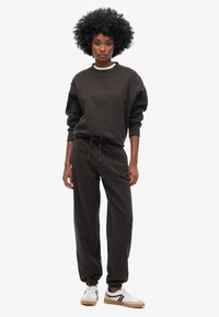 Donkerbruine sweatshirt en bijpassende joggers van zacht materiaal, met een ronde hals en elastische boorden. Witte accenten op de halslijn; sneakers met zwarte strepen.
