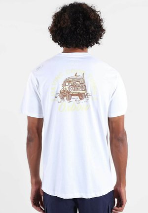 Homme aux cheveux bouclés portant un t-shirt blanc avec un motif représentant un véhicule tout-terrain avec un vélo et une planche de surf à l'arrière.