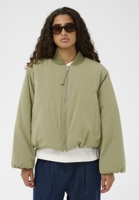 Svijetlo maslinasto zelena bomber jakna s patentnim zatvaračem, rebrastim ovratnikom i elastičnom stanicom. Ima glatku tkaninu i širok kroj.
