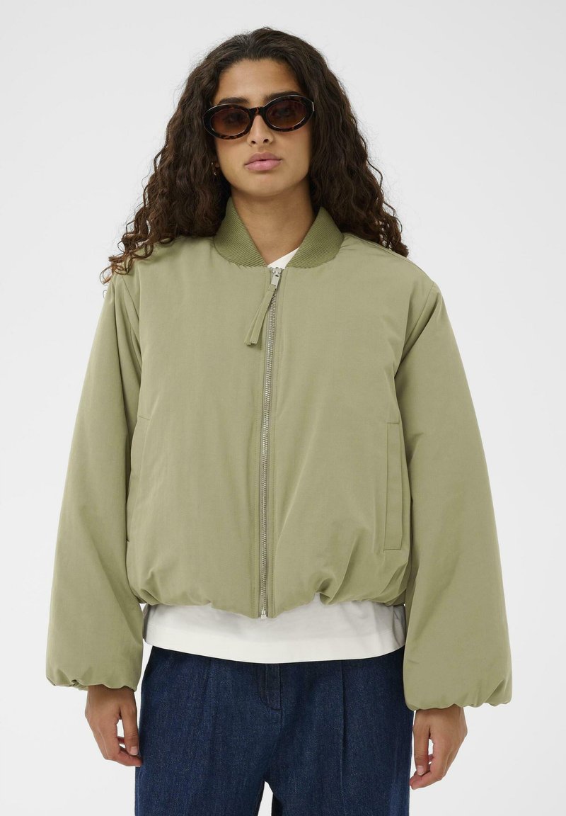 Svijetlo maslinasto zelena bomber jakna s patentnim zatvaračem, rebrastim ovratnikom i elastičnom stanicom. Ima glatku tkaninu i širok kroj.