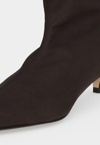 Gros plan d'une bottine en daim noir à bout pointu, avec un petit talon beige et des coutures visibles sur le côté.