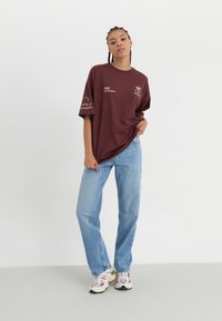 Burgunderfarget oversized t-skjorte med hvit tekst og palmetrær i grafikken på forsiden, kombinert med lyseblå rettlinjede jeans og flerfargede sneakers.