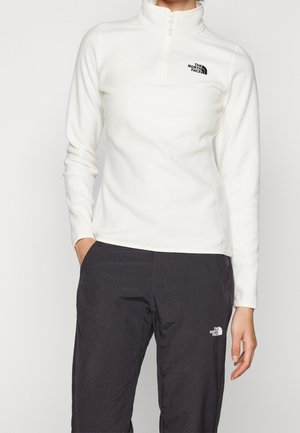 Persoon draagt een witte fleece trui met kwartrits en zwarte broek, beiden met het The North Face-logo, staand met handen deels in de zakken.
