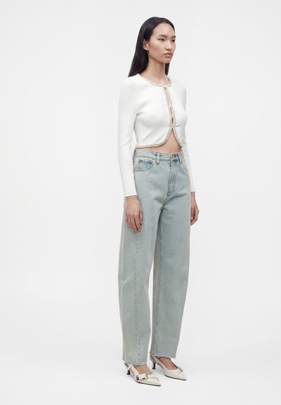 Femme portant un cardigan court blanc avec bordure en perles, un jean taille haute bleu clair et des chaussures crème à talons bas pointues, debout devant un fond blanc.