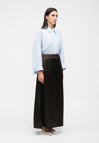 Γυναίκα με ίσια σκούρα μαλλιά, που φοράει ένα ανοιχτό μπλε oversized πουκάμισο, μακριά μαύρη φούστα και μπεζ μυτερά παπούτσια, στέκεται μπροστά σε απλό φόντο.