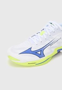 Zapato deportivo blanco con acentos azules, con una superficie texturizada, parte superior de malla y una suela verde brillante. Logotipo de la marca visible en el lateral.