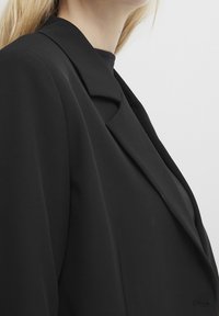 Blondynka w czarnym żakiecie na czarnej bluzce, sfotografowana od ramienia do podbródka na białym tle.