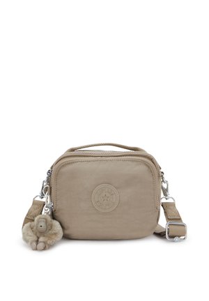 Bolso bandolera de tela beige con cierre de cremallera, superficie texturizada, logotipo circular y un llavero de mono peludo colocado en la correa.