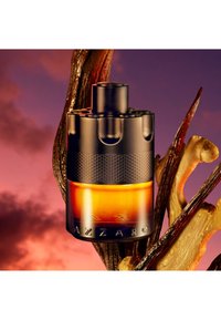 Zwarte en amberkleurige Azzaro parfumfles weergegeven met vanillestokjes en gemberwortels tegen een achtergrond van een paarse en oranje zonsondergang.