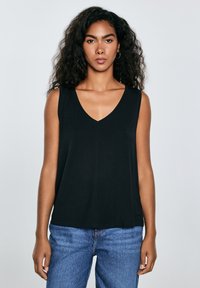 Mujer con cabello rizado y oscuro, vistiendo un top negro sin mangas con escote en V y jeans azules, de pie frente a un fondo gris claro.